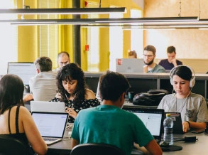 Pessoas reunidas em uma área comum da Aldeia Coworking Estação em Curitiba, sentadas em pufes e bancos de madeira, em um ambiente moderno e colaborativo com iluminação criativa e plantas decorativas.